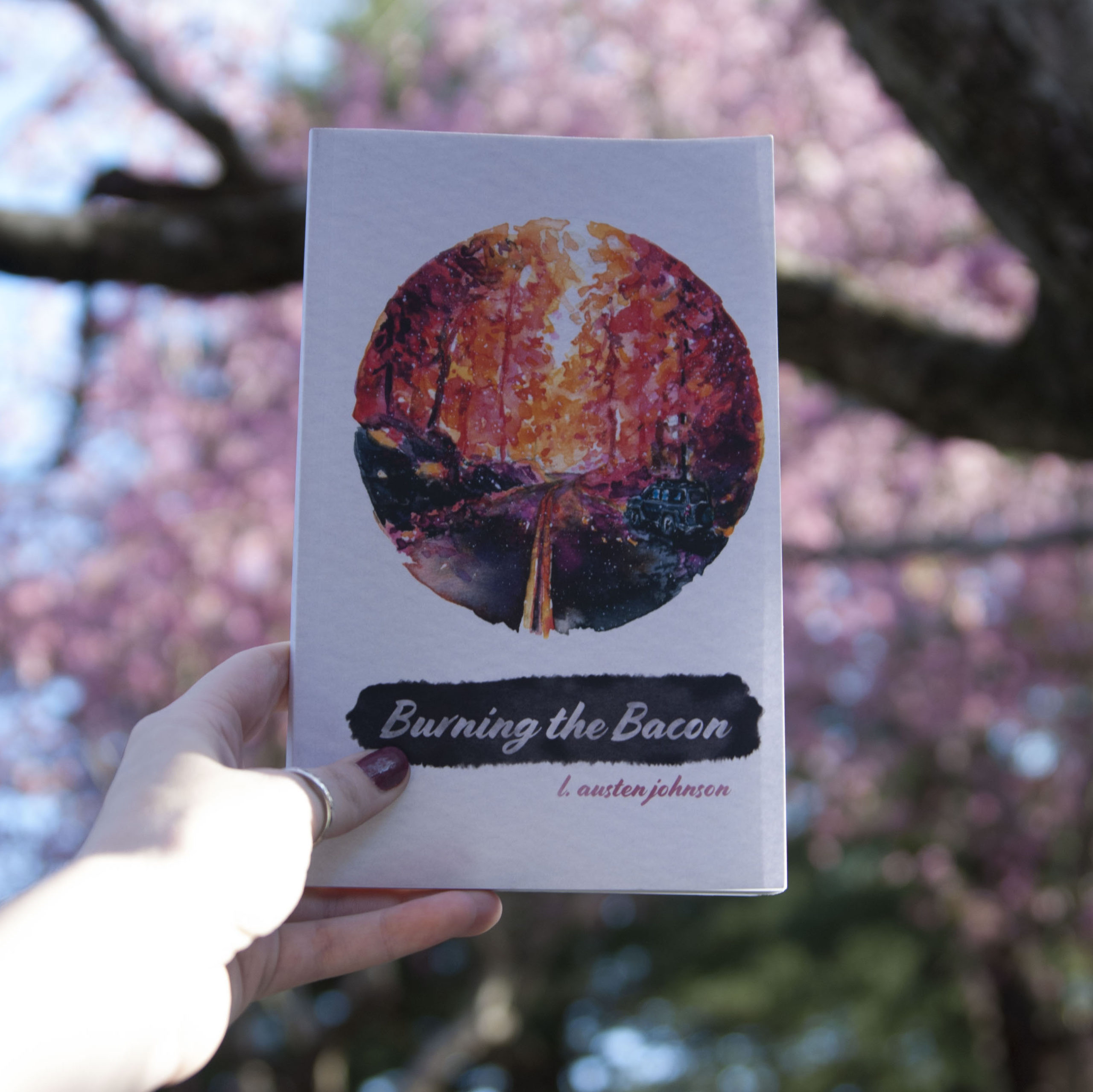 A hand holds the poetry collection Burning the Bacon in front of a pink cherry blossom tree